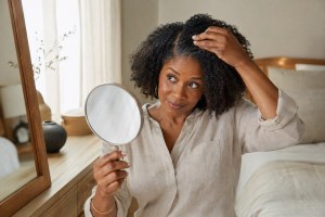 A woman with natural textured curls gently examines her hair and scalp in a hand mirror.