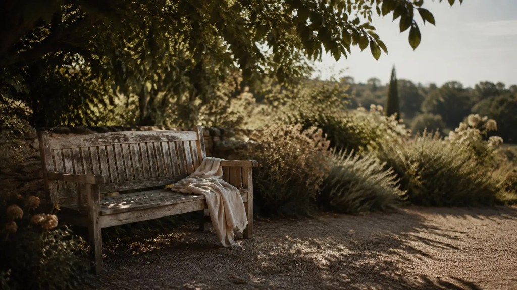 An empty wooden garden bench with an ivory linen throw beneath leafy late-summer shade.